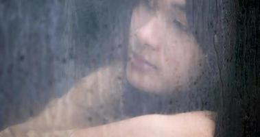 närbild skott, asiatisk ledsen ung kvinna tittar genom fönstret på regnig dag. depression koncept. video