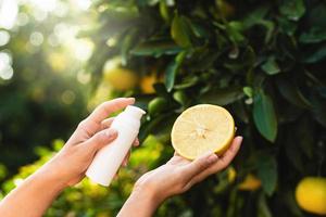 la mujer sostiene una botella de producto para el cuidado de la piel y la mitad de un limón en sus manos sobre el fondo del árbol de limón.