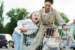feliz madre y sus hijas se divierten con un carrito de compras en un estacionamiento al lado de un supermercado.