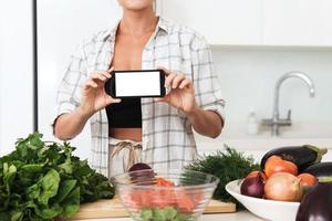 Woman is holding smartphone with blank screen for your design during cooking in modern kitchen photo