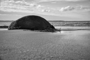 Big stone shot in black and white, on sandy beach in front of sea with clouds in sky photo