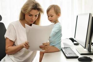 Young freelancer mother reading papers at home office with son sitting on desk. photo