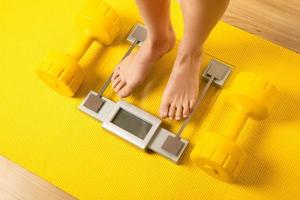 Female feet on the modern weighing scale with a yellow dumbbells and fitness mat photo
