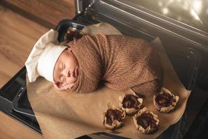 Newborn baby wearing chef's hat is lying on the oven tray with a muffins