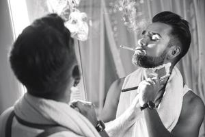 Man looking in the mirror, smoking a cigarette and shaving his beard with a straight razor photo