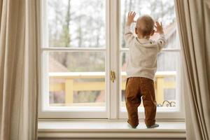 Baby boy is standing on the windowsill and looking at the window photo