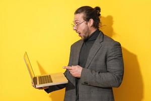Surprised man wearing eyeglasses pointing at laptop screen on yellow background photo