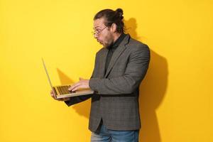 Surprised man wearing eyeglasses is using laptop computer on yellow background photo