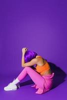 Young woman wearing colorful sportswear sitting against purple background photo