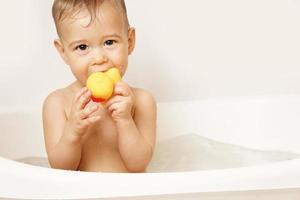 niño pequeño poniendo pato de goma en la boca mientras toma un baño. foto