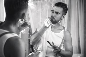 hombre mirándose en el espejo, fumando un cigarrillo y afeitándose la barba con una navaja de afeitar foto