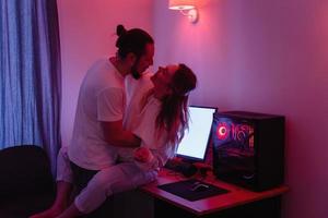 Cheerful couple in room with neon light, having fun on the table with gaming personal computer photo