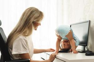 Little boy standing upside down on a desk and distracting mother from work on computer. photo