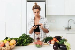 mujer con auriculares inalámbricos está usando un teléfono inteligente durante la cocina en una cocina blanca moderna foto
