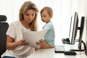 Young freelancer mother reading papers at home office with son sitting on desk. photo