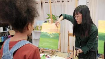 Female Asian teacher teaches and demonstrates to Black student girl on acrylic color picture painting on canvas in art classroom, creatively learning with skill at elementary school studio education. video