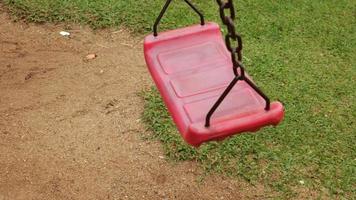 faites un ralenti le long d'une balançoire rouge attachée à une vieille chaîne rouillée dans la cour de récréation. dans le parc vu le sol et l'herbe. video