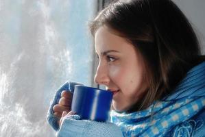 una mujer joven y triste que se queda en casa debido a la gripe y la temperatura en un suéter cálido y una bufanda con una taza de té cerca de la ventana foto