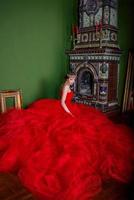 hermosa mujer con vestido rojo largo y corona real cerca de la chimenea en interiores de lujo en estudio fotográfico foto