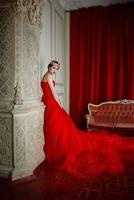 Beautiful woman in long red dress and in royal crown near fireplace in luxury interior in photo studio