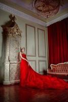 Beautiful woman in long red dress and in royal crown near fireplace in luxury interior in photo studio