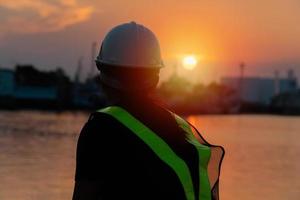 Silhouettes of asian woman engineer holding wrenches and radio communication walkie talkie she standing on shipyard. Background is oil storage silo. Concept of women girl power equal opportunity. photo