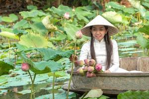 Beautiful asia women wearing white traditional Vietnam dress Ao Wai and Vietnam farmer's hat and sitting on wooden boat in flower lotus lake. Her hands picking lotus flowers. photo