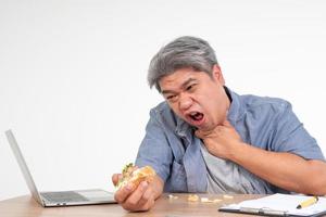 Asian man working and eating a burger on office desk and holding his neck after choking foods. Concept of a busy businessman cannot work-left balance and not taking care of health Eat only junk food photo