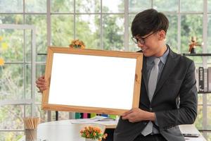 Happy Asian young businessman holding an empty white board and sitting on the table in office. Concept of copy space for advertisement, text, and content. photo