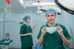 retrato de un cirujano asiático con mascarilla médica de pie en el quirófano de un hospital. equipo de cirujanos profesionales. salud, concepto de servicio médico de emergencia foto