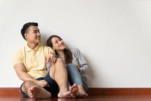 feliz pareja joven asiática sentada en el suelo en casa y mirando hacia arriba con espacio de copia apuntando hacia el aire en el fondo de la pared blanca foto