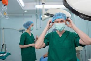 retrato de un cirujano asiático con mascarilla médica de pie en el quirófano de un hospital. equipo de cirujanos profesionales. salud, concepto de servicio médico de emergencia foto
