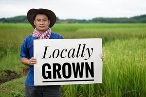 Asian man farmer holds paper banner with text  Locally Grown at paddy field. Concept, campaign to do organic agriculture,  locally grown style with no pesticide and herbicide or chemicals. photo