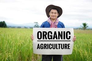 Asian man farmer holds paper banner with text  Organic Agriculture at paddy field. Concept, campaign to do organic agriculture, no pesticide and herbicide or chemicals. photo