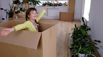 The girl hides in a big box and looks out of it funny in a room inside the home. Moving to a new house, unpacking things, renting an apartment, mortgage, housing issue video