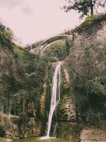 hermosa cascada de montaña con una corriente de agua que fluye desde debajo de un puente en un acantilado cubierto de árboles y plantas foto