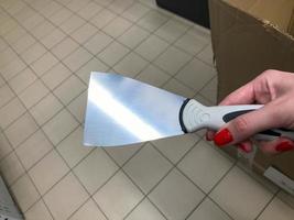 raspador para aplicar masilla, yeso. trabajos de pintura. un raspador en manos de una mujer con una manicura roja brillante. raspador con mango de plástico. contra el fondo de cajas en la tienda foto