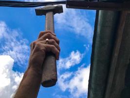a man hammers nails with a hammer against the blue sky. repairs the roof of a wooden house. the man is wearing a gold ring. holds a hammer, carries out repair work at a height photo