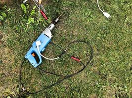 a drill with a long drill inside lies on the grass. renovation in nature. next to a hacksaw, hammer and other tools. the drill is connected through a white extension cord. building a village house photo