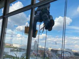 un trabajador de lavado de ventanas masculino, escalador industrial cuelga de un edificio alto, rascacielos y lava grandes ventanas de vidrio para limpieza en lo alto de una gran ciudad foto