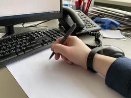 A man's hand in a shirt and with a fitness bracelet holds a pen and writes on the table at the office table with a computer with a keyboard. Business work photo