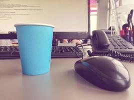 taza de café azul de cartón de una cafetería en un escritorio con una computadora con teclado y mouse y suministros de oficina de un trabajador de oficina durante el almuerzo. pausa para el café de negocios foto