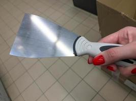 scraper for applying mastic, plaster. Painting works. DIY home repair. a scraper in the hands of a woman with a bright red manicure. scraper with metal tip and plastic handle photo