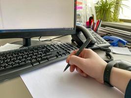 A man's hand in a shirt and with a fitness bracelet holds a pen and writes on the table at the office table with a computer with a keyboard. Business work photo