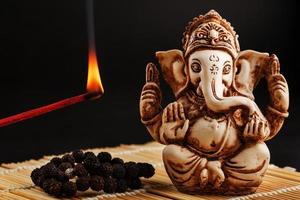 Hindu god Ganesh on a black background. Rudraksha statue and rosary on a wooden table with a red incense stick and incense smoke photo