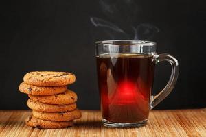 A pile of oatmeal cookies with chocolate chips and a mug of fragrant black hot tea in on a bamboo substrate, on a dark background. Handmade cookies for a healthy breakfast photo