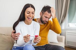 Not ready, worried man checking a pregnancy test with his excited wife sitting on a couch at home photo