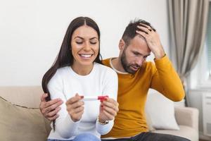 Not ready, worried man checking a pregnancy test with his excited wife sitting on a couch at home photo