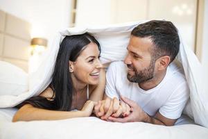 smiling couple going to kiss under sleeping sheets in the morning. In love young happy couple lying in bed in the morning photo