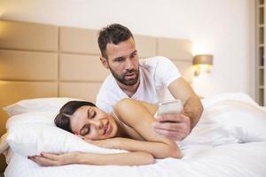 Jealous husband spying the phone of his partner while she is sleeping in a bed at home. Shocked jealous husband spying the phone of his wife while she sleeping in bed at home photo